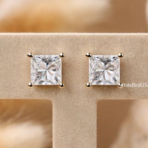 May include: A pair of square-cut, clear gemstone stud earrings with gold-colored settings. The earrings are displayed on a beige stand against a blurred background of dried plants and a light-colored stone.