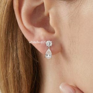 May include: A close-up of a person's ear adorned with silver drop earrings. Each earring has a round, clear stone at the top and a pear-shaped stone suspended below. The earrings are set against the person's skin. The earrings sparkle.