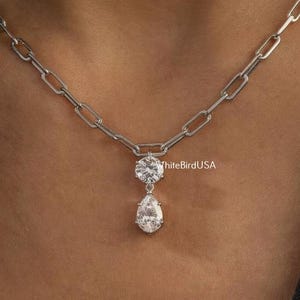May include: A silver chain necklace with a double-pendant design. The top pendant is a round, clear gemstone, and the bottom pendant is a pear-shaped, clear gemstone. The necklace is worn on a person with dark hair and a black shirt.