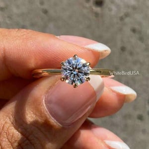 May include: A gold engagement ring featuring a round, clear gemstone. The ring has a simple, elegant design with a slender band. The gemstone is secured by six prongs. The ring is set against a grey background.