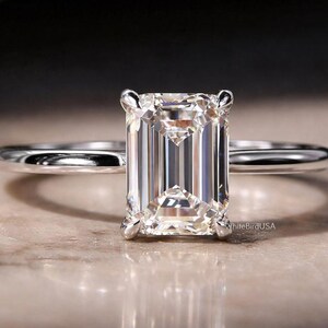 May include: A solitaire engagement ring with an emerald-cut diamond. The diamond is set on a thin, polished silver band. The ring is displayed on a neutral-toned surface, with the brand name "White Bird USA" visible.