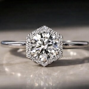 May include: A silver engagement ring with a hexagonal halo of small diamonds surrounding a large, round, clear center stone. The ring is set on a thin band and sits on a light-colored surface.