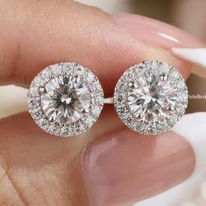 May include: Two round diamond stud earrings with a halo of smaller diamonds. The earrings are held up by a hand with a light pink manicure. The earrings are set in a silver metal. The WhiteBirdUSA logo is visible in the background.