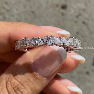 May include: A silver ring with a continuous row of clear, marquise-cut stones. The ring is held against a gray background, showcasing the sparkle of the gemstones. The ring is a piece of jewelry.