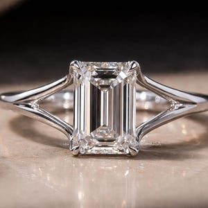 May include: A silver ring featuring an emerald-cut diamond. The diamond is set in a split shank design. The ring is displayed on a neutral surface. The ring is a piece of jewelry.