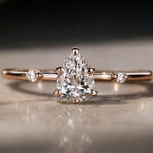 May include: A rose gold engagement ring with a pear-shaped diamond center stone. The ring features two smaller round diamonds on either side of the center stone. The band is thin and delicate, with a polished finish. The ring is set on a reflective surface.
