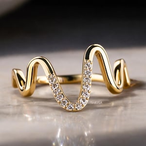 May include: A gold-colored ring with a wavy design. The ring features a series of small, clear stones set in a curved section, creating a sparkling accent. The ring's design is modern and elegant, suitable for everyday wear or special occasions.