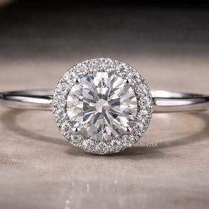 May include: A silver engagement ring with a large, round, brilliant-cut diamond center stone. The center stone is surrounded by a halo of smaller diamonds. The ring is set on a thin, polished band. The ring is a classic and elegant design.