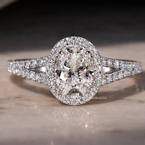May include: An oval-cut diamond engagement ring with a halo of smaller diamonds. The ring features a split shank band, also encrusted with diamonds. The ring is set on a neutral-toned surface, with soft lighting.