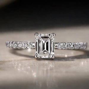 May include: A close-up of a diamond ring with an emerald-cut center stone. The band is adorned with smaller diamonds. The ring is set on a neutral surface, with soft lighting.