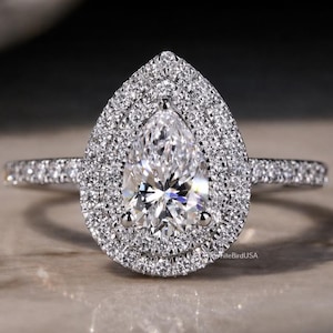 May include: A pear-shaped diamond ring with a double halo of smaller diamonds. The ring's band is also encrusted with diamonds, creating a sparkling effect. The ring is set on a neutral surface, reflecting the light.