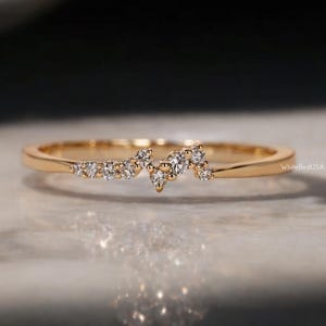 May include: A slender gold ring featuring a cluster of small, sparkling diamonds. The diamonds are arranged in an unusual, asymmetrical design. The ring sits on a reflective surface, with the text "WhiteBirdUSA" visible.
