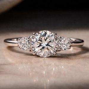 May include: A three-stone engagement ring with a large, round, brilliant-cut diamond center stone and two smaller side stones. The ring is set in a silver band and sparkles brightly. The ring is on a light-colored surface.
