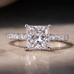 May include: A close-up of a diamond engagement ring. The ring features a large, square-cut diamond as the centerpiece, with smaller diamonds set along the band. The ring is set in a silver or white gold band.