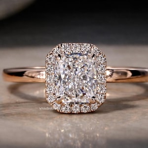 May include: Rose gold engagement ring featuring a radiant-cut diamond center stone surrounded by a halo of smaller diamonds. The ring has a delicate band and a classic, elegant design, perfect for a proposal or special occasion.