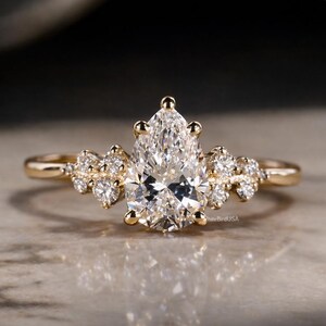 May include: A gold ring featuring a pear-shaped diamond center stone, accented by smaller diamonds on the band. The ring has a delicate, elegant design, perfect for engagement or special occasions. The ring is set on a reflective surface.