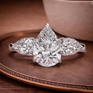 May include: A platinum ring with a large, pear-cut diamond centre stone, accompanied by two smaller pear-cut diamonds. The ring's design is elegant and classic, highlighting the diamonds' brilliance. The ring is set on a brown surface.