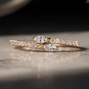 May include: A gold ring with a bypass design. The ring features two marquise-cut diamonds and is embellished with smaller diamonds along the band. The ring is set on a light-coloured surface.