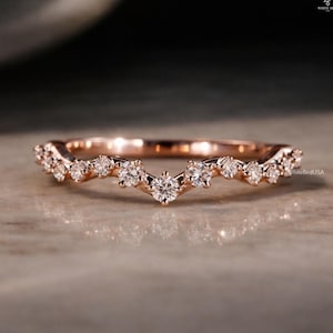 May include: Rose gold band ring with a curved design, set with multiple small, clear gemstones. The ring has a delicate, elegant appearance, perfect for a special occasion. The ring is on a neutral surface.