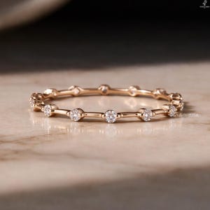 May include: A delicate rose gold ring with small, round, clear gemstones set along the band. The ring is displayed on a light-colored, textured surface. The ring is a piece of jewelry.