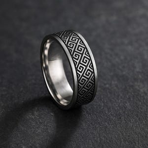 Könnte beinhalten: Ein silberner Ring mit einem geometrischen Muster, das um das Band geätzt ist. Der Ring hat eine polierte Oberfläche und ein dunkelgraues Muster. Der Ring ist vor einem dunklen Hintergrund platziert.