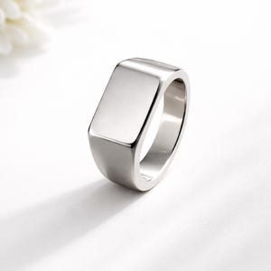 May include: A polished silver ring with a rectangular face. The ring has a smooth, reflective surface and a rounded band. The jewellery is set against a white background, highlighting its minimalist design.