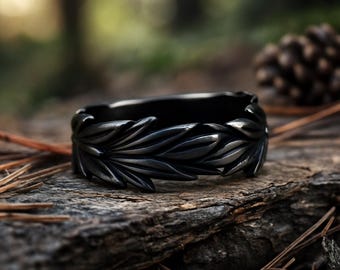 May include: A dark gray ring with a leaf design. The ring is made of metal and has a detailed pattern of leaves around the band. The ring is set against a natural background of wood and pine needles.