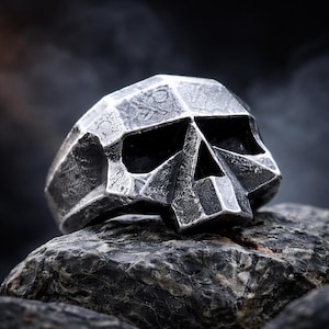 May include: A silver-colored skull ring with a geometric design. The ring features a detailed, textured surface and is set against a dark, blurred background. The skull's eye sockets and nose cavity are visible.