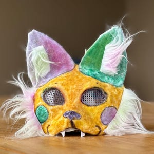 Puede incluir: Una máscara de gato hecha a mano con detalles en amarillo, verde y morado. La máscara presenta orejas grandes y puntiagudas y está adornada con suave pelo sintético blanco y rosa. Los ojos están cubiertos con un material de malla y la máscara tiene pequeños dientes.