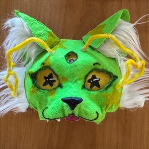 May include: A handmade green and yellow creature mask with white fur accents and yellow pipe cleaner antennae. The mask features star-shaped eyes and a black nose, perfect for costume parties or theatrical performances.