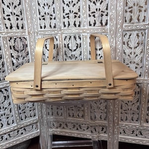 May include: A light-colored, woven picnic basket with a wooden lid and two arched handles. The basket is rectangular with rounded edges and a hinged lid. The handles are made of bent wood. The basket is sitting on a clear surface.
