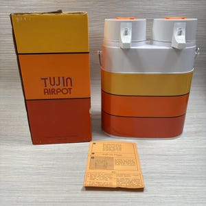 Puede incluir: Un Airpot Tujin vintage con su caja original y hoja de instrucciones. El airpot es blanco, amarillo y naranja con dos boquillas y un asa. La caja es naranja y amarilla con las palabras "Tujin Airpot" impresas.