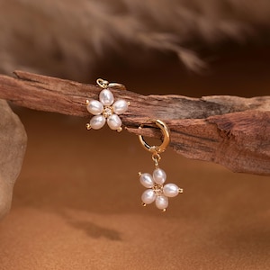May include: Gold-tone flower earrings with white pearl petals and a small crystal center. One earring is a stud, and the other is a hoop with a dangling flower. The earrings are displayed on a piece of wood.