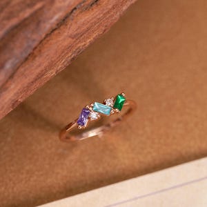 May include: A gold-toned ring featuring a cluster of three baguette-cut gemstones in purple, light blue, and green, accented by two small clear round stones set along a thin, polished metal band that encircles the finger.