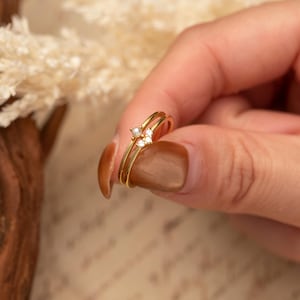 Puede incluir: Un delicado juego de anillos de oro con un diseño de doble banda. La banda superior está adornada con una pequeña perla y dos piedras preciosas transparentes, creando un aspecto sutil y elegante. El anillo se sostiene sobre un fondo suave y neutro.