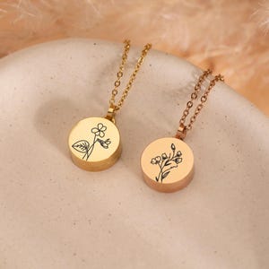 May include: Two gold-toned necklaces featuring circular pendants. Each pendant is engraved with a distinct black line-art floral design. The necklaces have delicate link chains and a polished metallic finish, showcasing a simple and clean aesthetic.