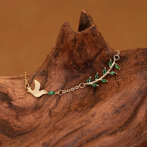 May include: A gold-colored necklace featuring a dove and a branch with green gemstones. The necklace is delicate and has a chain link design. The dove is on the left side of the necklace.