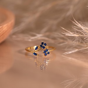 Può includere: Un anello color oro caratterizzato da un delicato design a ramo, impreziosito da piccole pietre preziose blu scuro disposte in motivi floreali e a foglia. La fascia è sottile e lucida, con una superficie riflettente dal design chiaro e simmetrico.