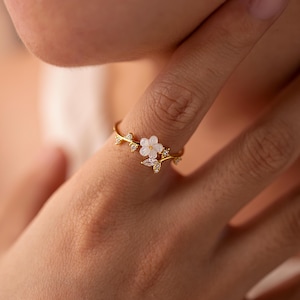 Peut inclure: Bague dorée ornée d'un motif floral délicat. La bague présente une petite fleur blanche avec des accents de pierres précieuses claires et des détails de feuilles. La bague est portée au doigt.