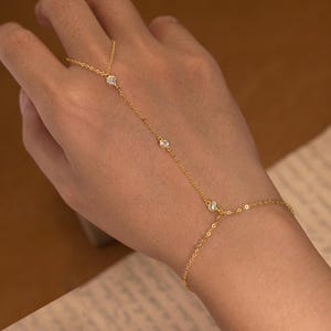 Pode incluir: Uma delicada corrente de mão dourada com três pedras preciosas redondas e transparentes. A corrente conecta-se a um anel e envolve o pulso. A joia é exibida em uma mão sobre um fundo neutro.