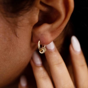 May include: A small gold hoop earring featuring a single dangling charm. The charm is a circular gold bead encrusted with tiny, clear, sparkling stones that catch the light. The hoop is a simple, polished gold band that secures through the earlobe.