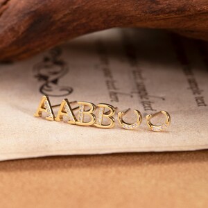 May include: Gold-colored stud earrings spelling out "AABBOO" with small, clear gemstones. The earrings are displayed on a vintage paper background with a wooden element in the upper portion of the image.