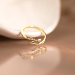 Può includere: Un anello aperto color oro caratterizzato da un design bypass con due estremità sovrapposte. La fascia è impreziosita da una fila di piccole pietre chiare e scintillanti incastonate lungo le curve frontali del metallo, creando un aspetto delicato.
