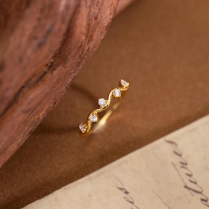 May include: A gold-colored ring with a wavy design, set with small, clear gemstones. The ring is displayed on a brown surface, with a wooden element in the background. The gemstones are evenly spaced along the ring's curve.