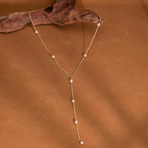 Puede incluir: Delicado collar dorado con pequeñas perlas espaciadas. El collar tiene un diseño en forma de Y, con una hebra más larga de perlas colgando. El fondo es marrón cálido. Una joya elegante y atemporal.