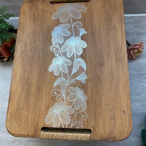 May include: A rectangular wooden serving tray with a floral design. The tray is made of light brown wood and features a vertical line of white flowers. The handles are wrapped with natural-colored rope.
