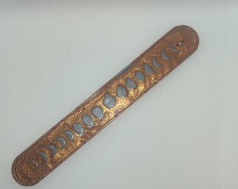 Puede incluir: Un porta incienso de color cobre con una serie de diseños de fases lunares plateadas. El soporte rectangular tiene extremos redondeados y un pequeño agujero en un extremo. Las fases lunares están dispuestas en una fila a lo largo del soporte.