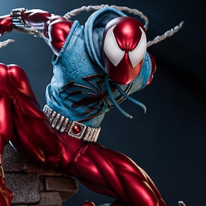 May include: A dynamic statue of a superhero in a red and blue costume. The figure is posed mid-action, with a red mask featuring white eye shapes. The costume includes a blue denim-style vest with a black spider emblem and a silver belt.