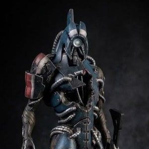 May include: A detailed figurine of a science fiction character in a blue and gray armored suit, holding a rifle. The figure has intricate mechanical details, standing on a textured base. The overall design suggests a futuristic or space-themed aesthetic.