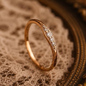 May include: A gold ring with a curved design, set with small, clear gemstones. The ring is displayed on a textured, off-white surface, with a decorative, ornate frame in the background. The ring's band has a hammered texture.